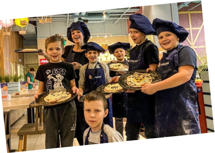 Děti v kuchařských čepicích a zástěrách drží podnosy s připravenými pizzami