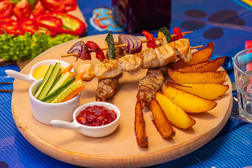 Masové špízy s grilovanou zeleninou, opečenými bramborami, zeleninovými tyčinkami a omáčkami na dřevěném prkénku